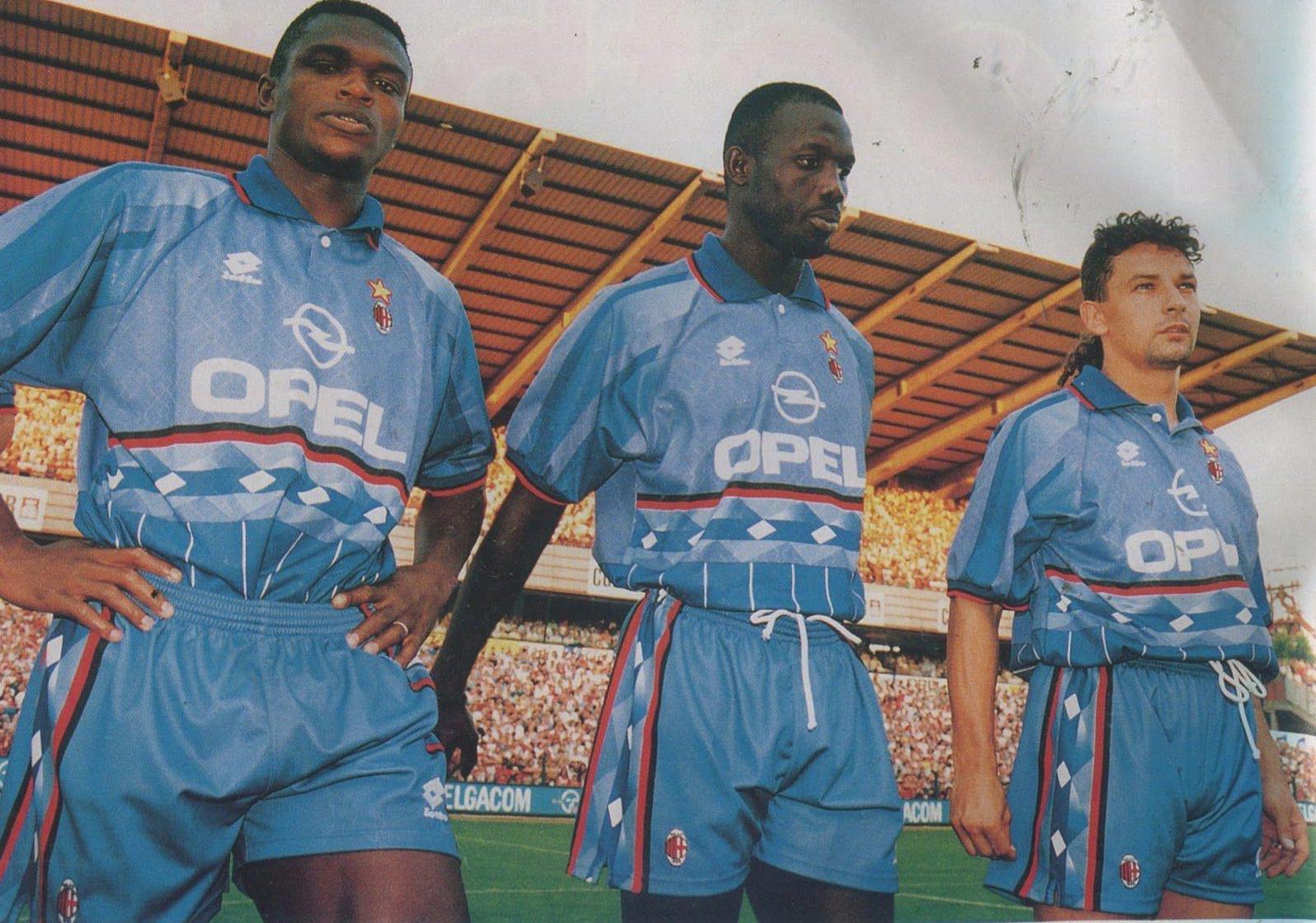 Three players in blue Retro1999 AC Milan 1995-96 Third Kit jerseys stand side by side on the field, serious and focused. The crowd and stadium seating form the backdrop for this authentic design with Opel as the sponsor.