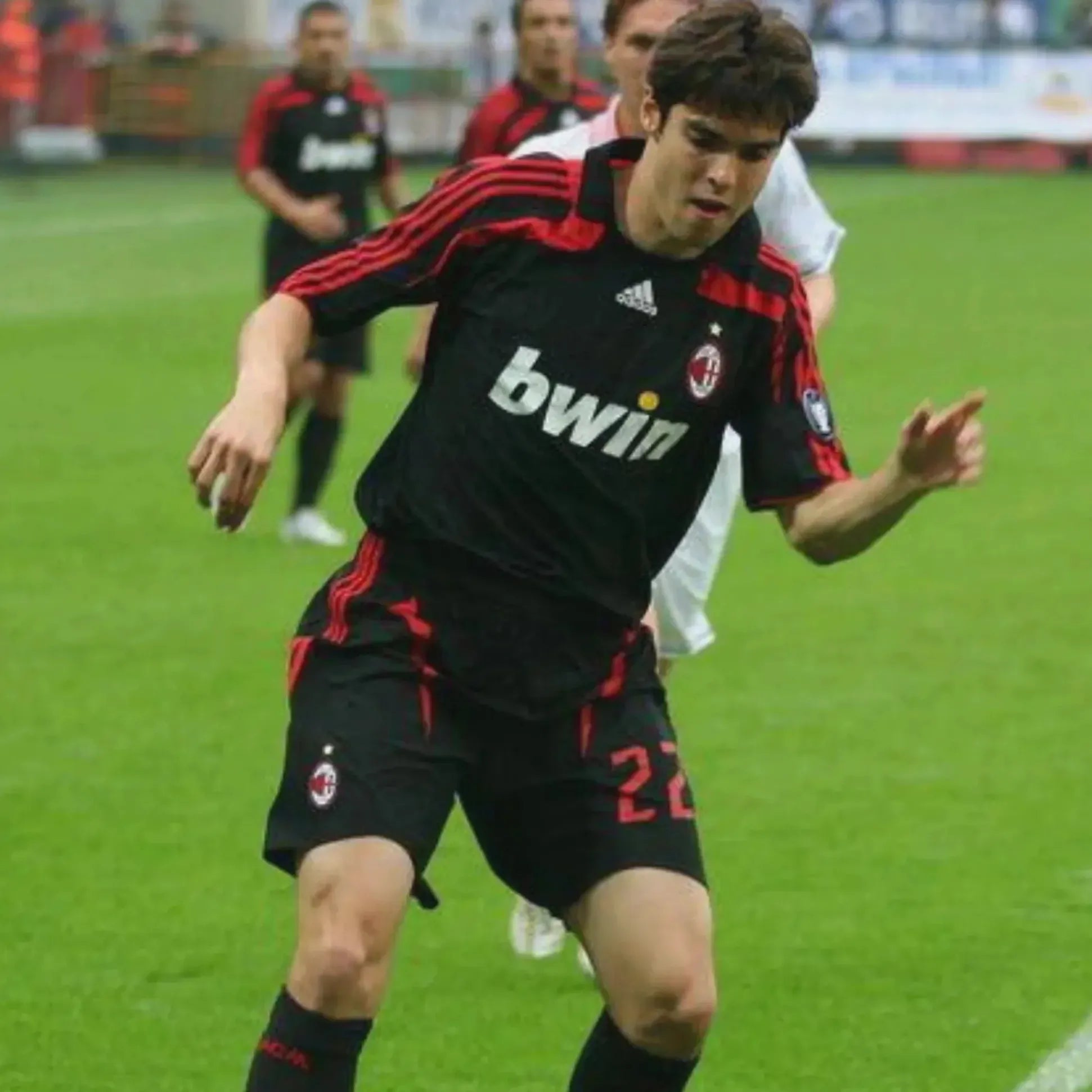 A soccer player in the Retro1999 AC Milan 2007-08 Third Shirt with number 22 on his shorts runs on a grassy field during a match, with other players and part of the audience seen in the background.