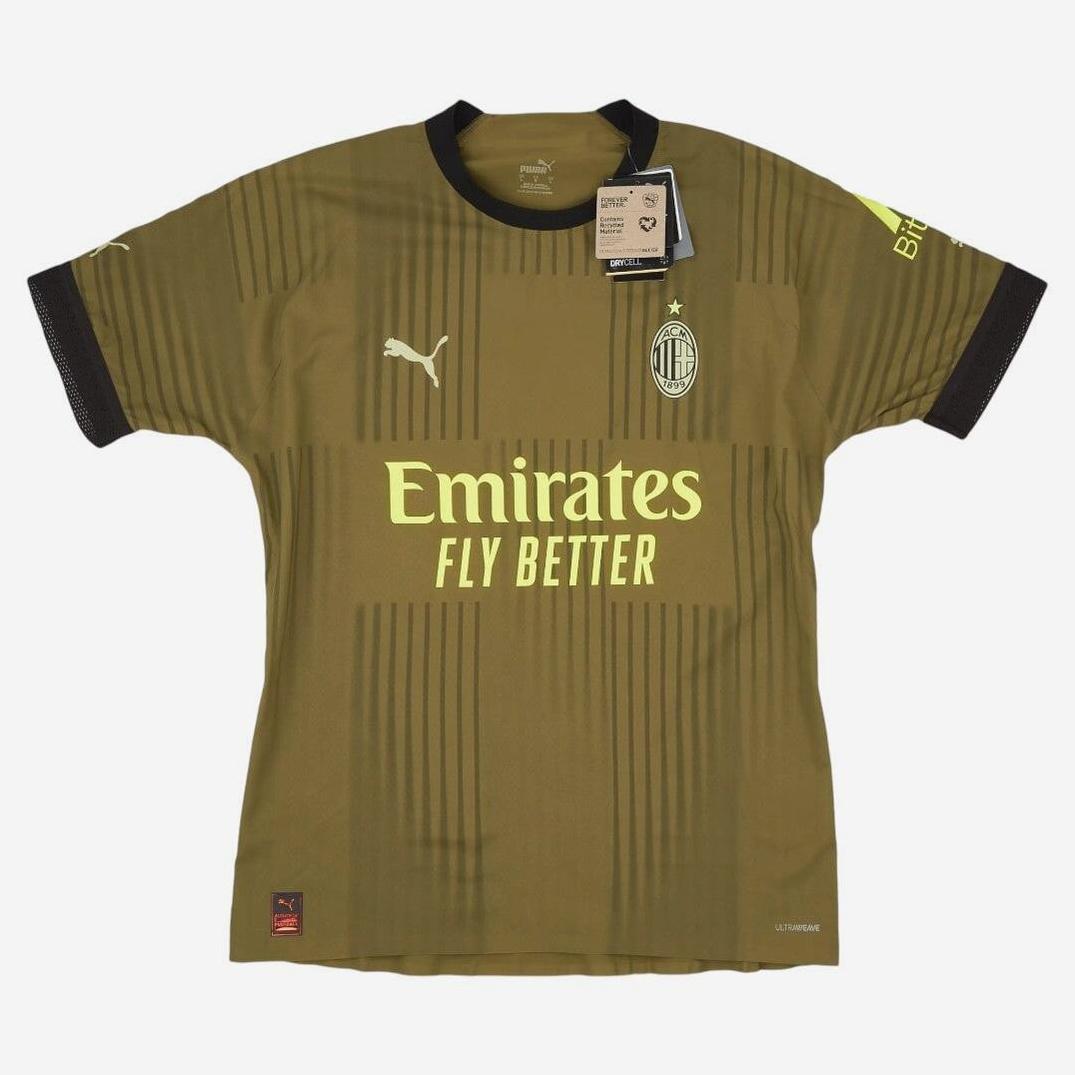 The AC Milan 2022-23 Third Shirt by Retro1999 features a gold retro-inspired style with black collar and sleeve cuffs, club crest on the left chest, central logos, and tags attached near the collar.