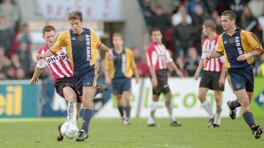 Two soccer players in action: one kicks the ball wearing the Retro1999 Ajax 2000-01 Away Shirt, while another in a blue-and-yellow kit runs beside him. Ajax fans and spectators are visible in the background.