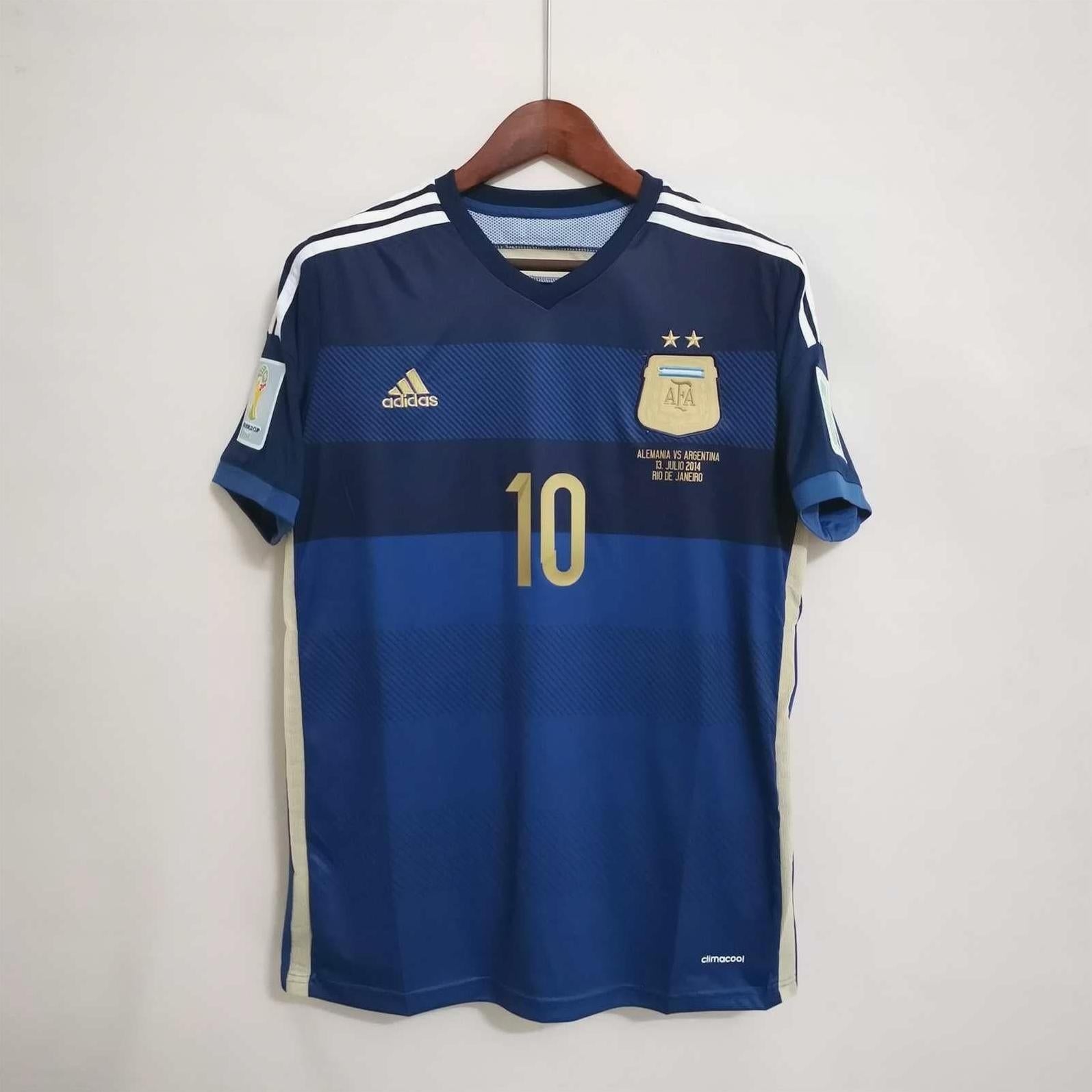 The Retro1999 Argentina 2014 Away Shirt in blue features the number 10, two gold stars above the crest, white shoulder stripes, and a retro design, hanging on a wooden hanger against a plain white wall.