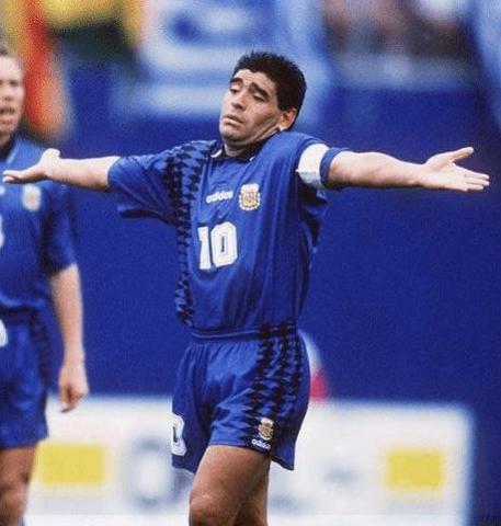 A soccer player in a blue Retro1999 Argentina Retro 1994 Away Shirt with number 10 stands triumphantly on the field, celebrating football history from World Cup ‘94, while another player is partially seen in the background.