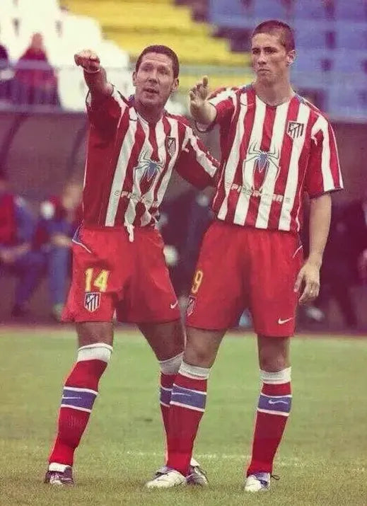 Two soccer players wear the Retro1999 Atletico Madrid 2004-05 Home Shirt, featuring iconic red and white stripes with the Columbia Pictures sponsor—one points ahead while the other gestures during a match.