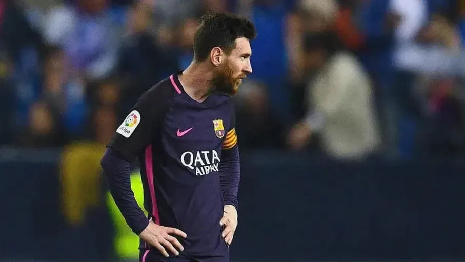 A footballer in a Retro1999 Barcelona 2016-17 Away Shirt with a captains armband stands on the field, hands on hips, facing sideways as fans in the blurred background recall an iconic Champions League comeback.