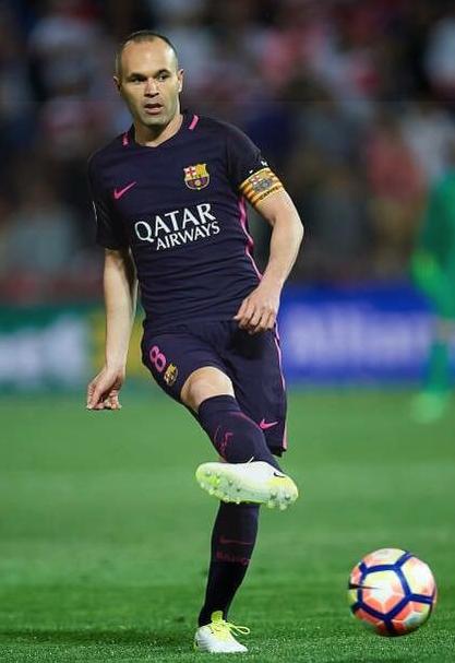 A soccer player in a Retro1999 Barcelona 2016-17 Away Shirt kicks a colorful ball on the field, wearing a captain’s armband and yellow shoes, standing out amid the blurred crowd during an exciting Champions League comeback.