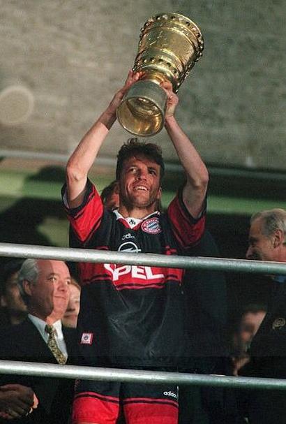 A soccer player in a Retro1999 Bayern Munich 1997-98 Away Shirt lifts a golden trophy, smiling as people in suits celebrate nearby, all surrounded by Bundesliga memorabilia.