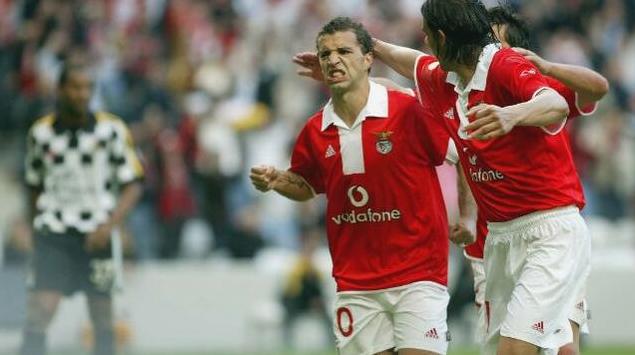 Two soccer players in red jerseys, sporting the Retro1999 Benfica 2004-05 Home Shirt, celebrate with passion on the field. In the background, a player in a black-and-white retro kit is blurred as fans cheer from the stands.