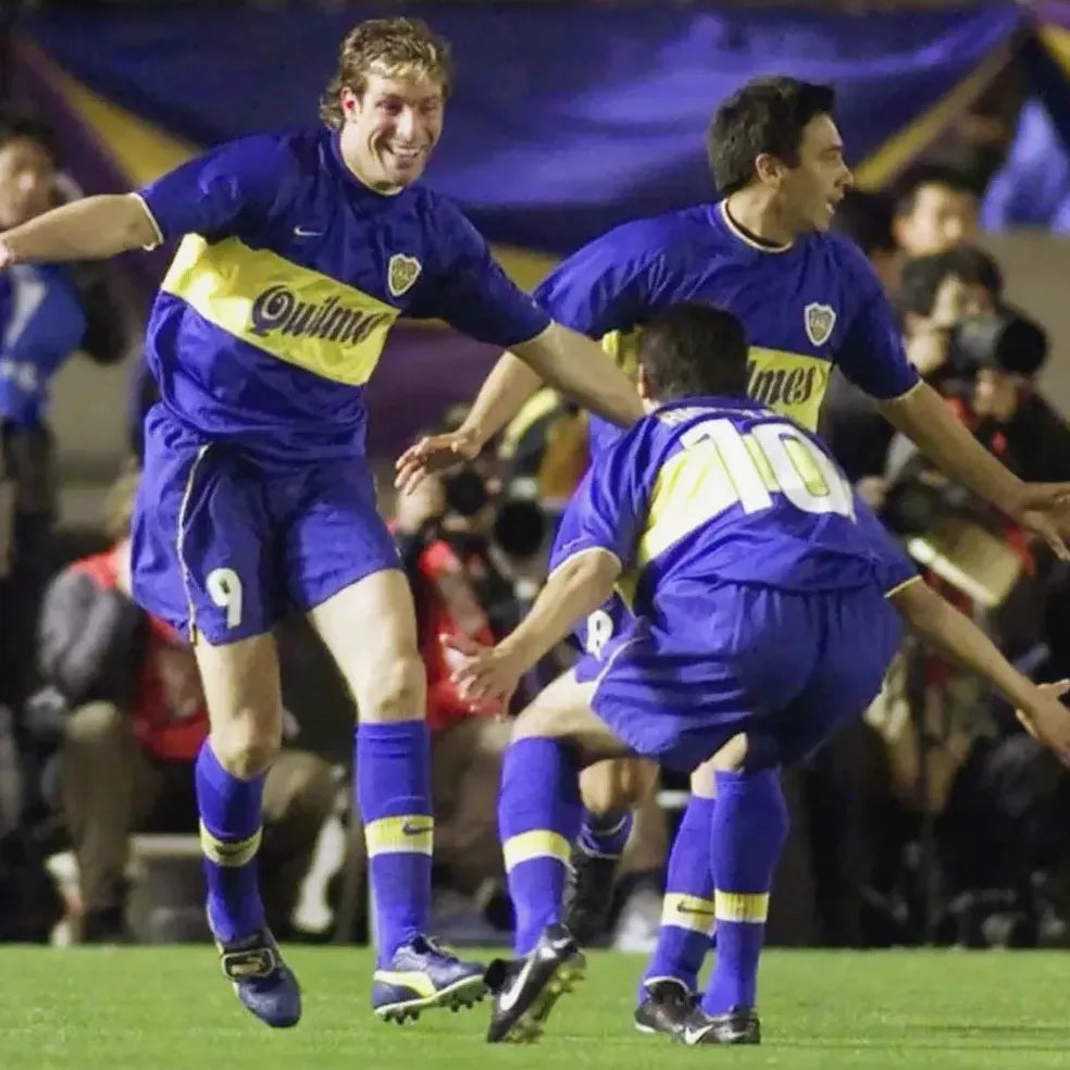 Three players in Boca Juniors 1999-00 Home Shirts by Retro1999 celebrate on the field, one smiling with arms outstretched, as photographers and fans capture a football moment to remember.