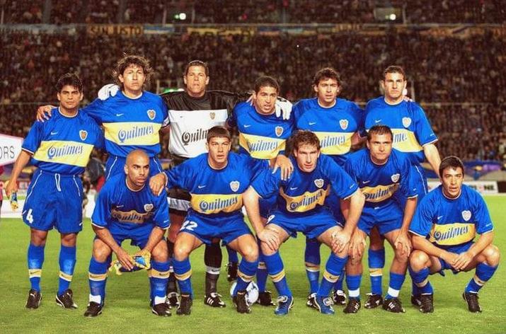 A soccer team in blue uniforms poses for a group photo on the field, wearing the Retro1999 Boca Juniors 1999-00 Home Shirt, with some players kneeling and others standing as fans fill the stadium in the background.