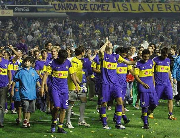 Boca Juniors players in the Retro1999 Boca Juniors 2010-11 Home Shirt, in purple and yellow, walk onto the field surrounded by fans and confetti, with a packed stadium in the background.