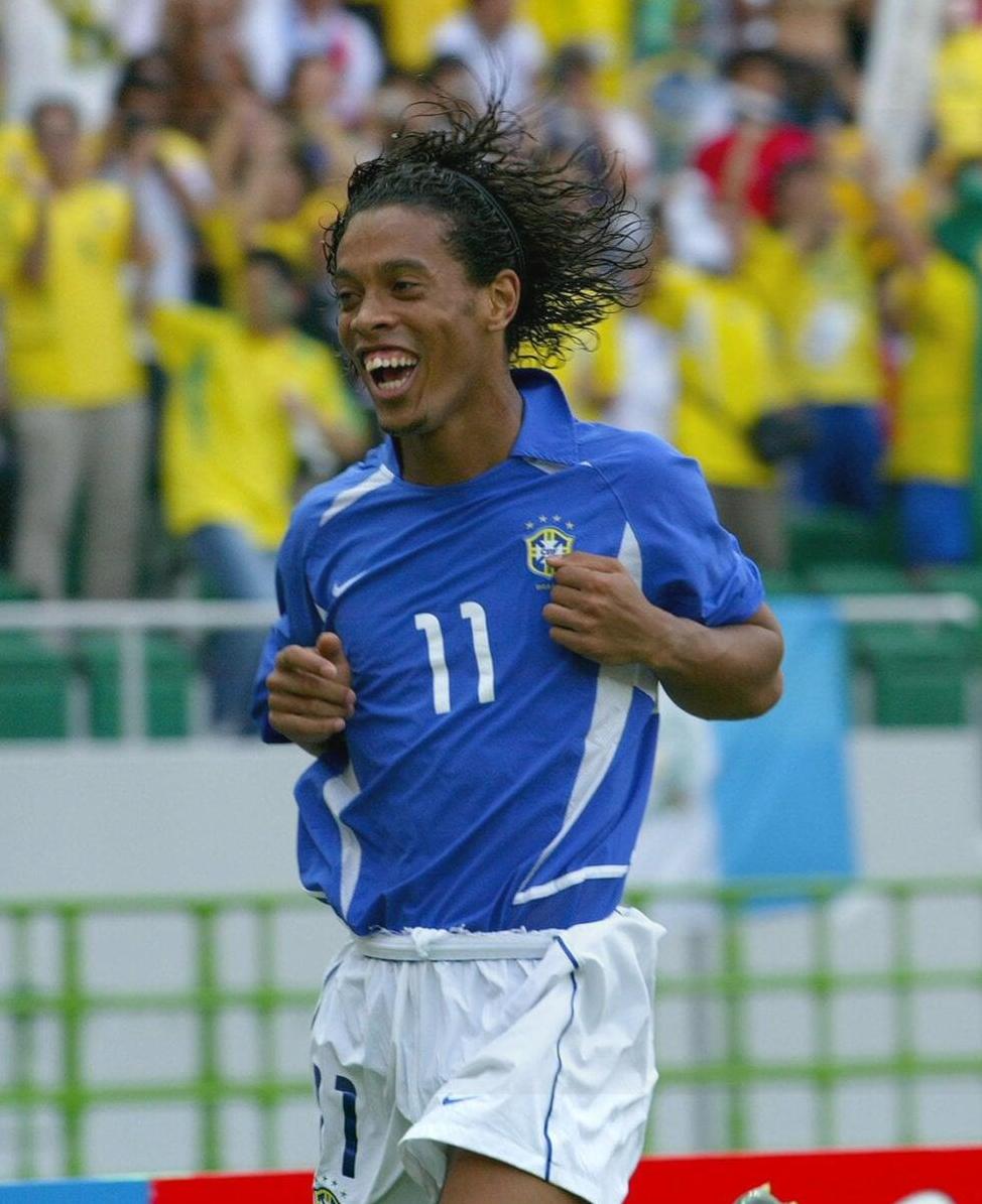 A soccer player in the Retro1999 Brazil 2002 Away Shirt, number 11, celebrates on the field with a bright smile and flowing hair. Fans in yellow cheer in the background, highlighting this classic World Cup scene.