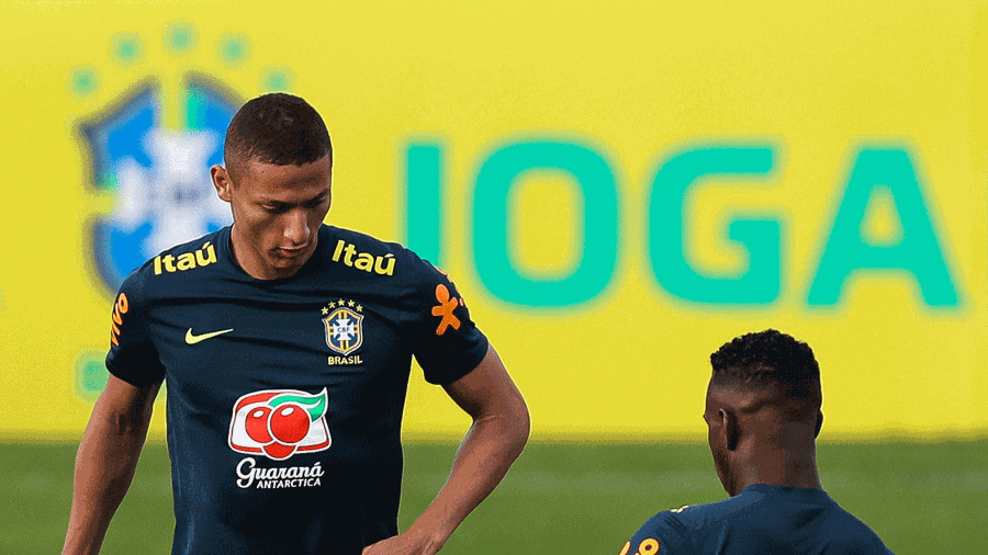 Two Brazilian national team players wear Retro1999 Brazil 2019 Training Shirts, standing before a vivid yellow backdrop with a blurred Brazil crest and JOGA in the background.