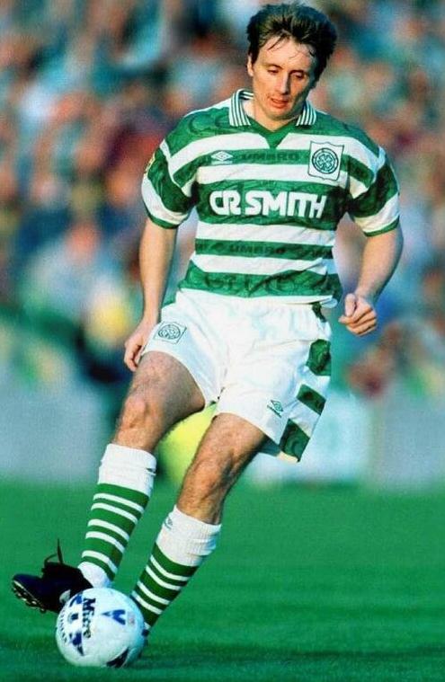 A soccer player wears the Retro1999 Celtic 1995-97 Home Shirt, showcasing green and white stripes with the CR SMITH logo, as he controls the ball on a grassy field in front of a blurred crowd.