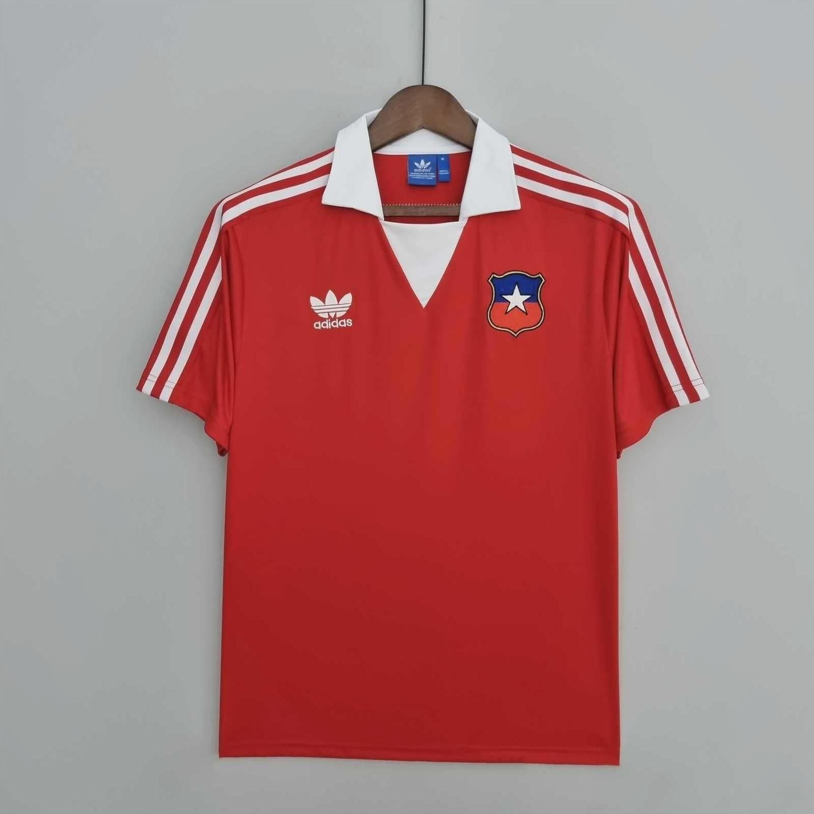 The Retro1999 Chile 1982 Home Shirt is a red short-sleeve soccer jersey with white shoulder stripes and a classic badge, displayed on a wooden hanger against a gray wall—a must-have retro kit for any Chile fan.