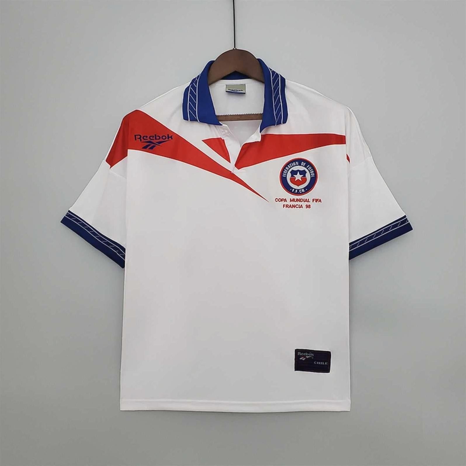 The Chile 1998 Away Shirt by Retro1999 is a white short-sleeve soccer jersey with blue collar and cuffs, red and blue details, plus a Reebok logo and the Chile football badge, inspired by the iconic Copa Mundial FIFA Francia 98 kit.