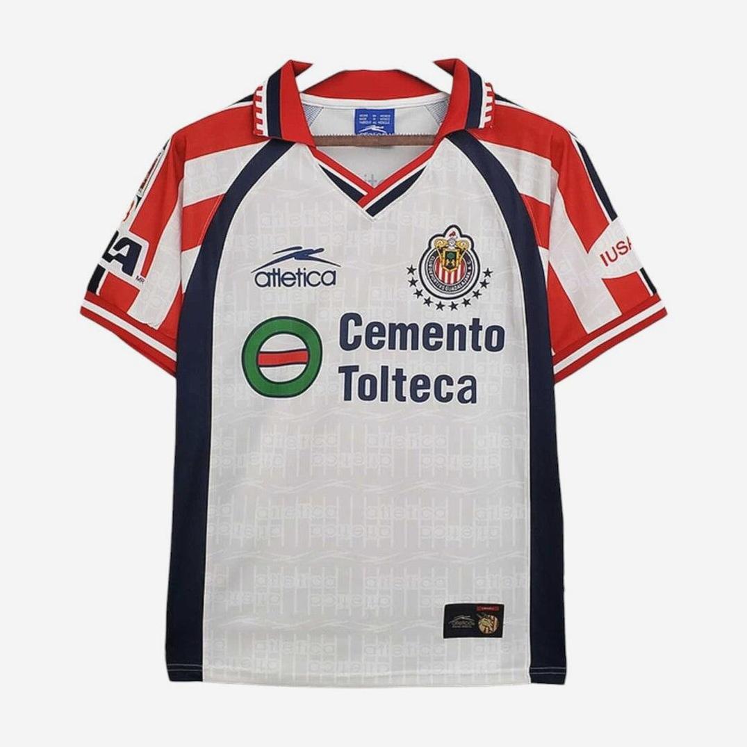 The Retro1999 Chivas 1999-00 Away Shirt features red, white, and blue stripes, team crest, Cemento Tolteca sponsor, and classic logos. A vintage kit with a blue v-neck collar and red-white sleeve accents—ideal for collectors.