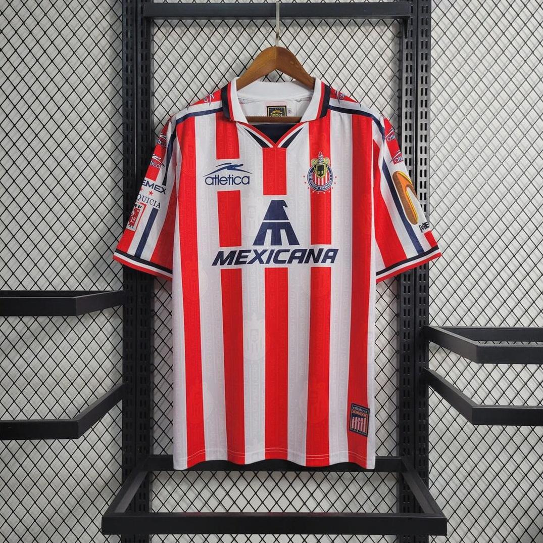 The Retro1999 Chivas 2006-07 Home Shirt—a red and white striped soccer jersey with blue accents—hangs on a black hanger against a mesh grid wall, featuring the Chivas logo and MEXICANA sponsor on the front. Perfect for fans!.