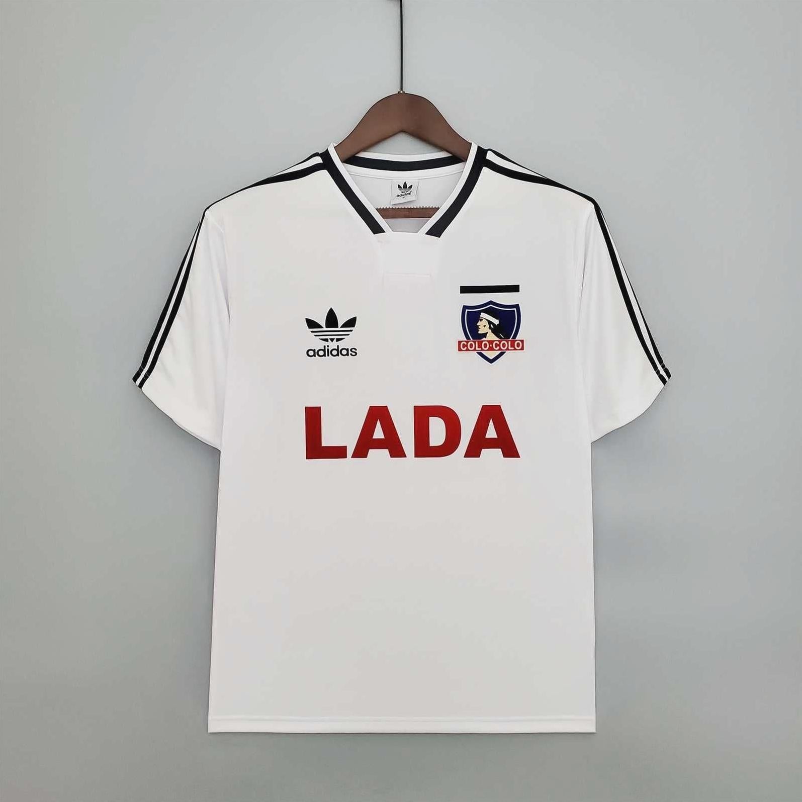 The Colo Colo 1991 Home Shirt by Retro1999 features a classic white design with black shoulder stripes, red LADA sponsor, and club crest on the upper left—ideal for vintage sports fans seeking an iconic retro kit. Displayed on a wooden hanger.