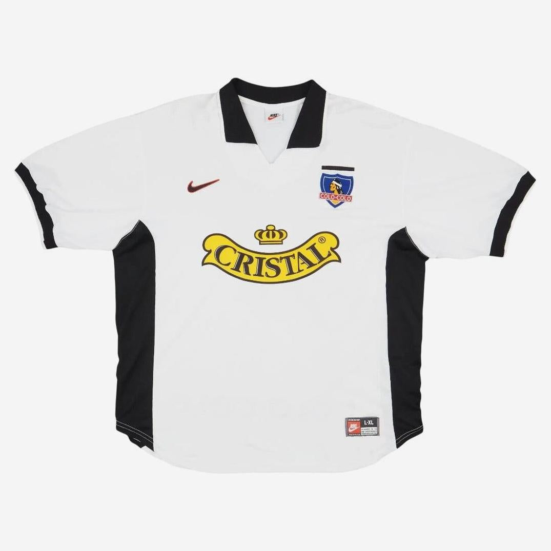 The Colo Colo 1998-99 Home Shirt by Retro1999 is a white jersey with black accents, black collar, club crest, Nike logo, and a bold yellow Cristal sponsor—ideal for football memorabilia collectors.
