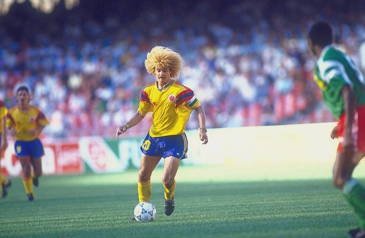A soccer player wearing the Retro1999 Colombia 1990 Home Shirt with number 10 and a large blond afro dribbles on the field, as teammates and a lively crowd fill the stadium in the background.