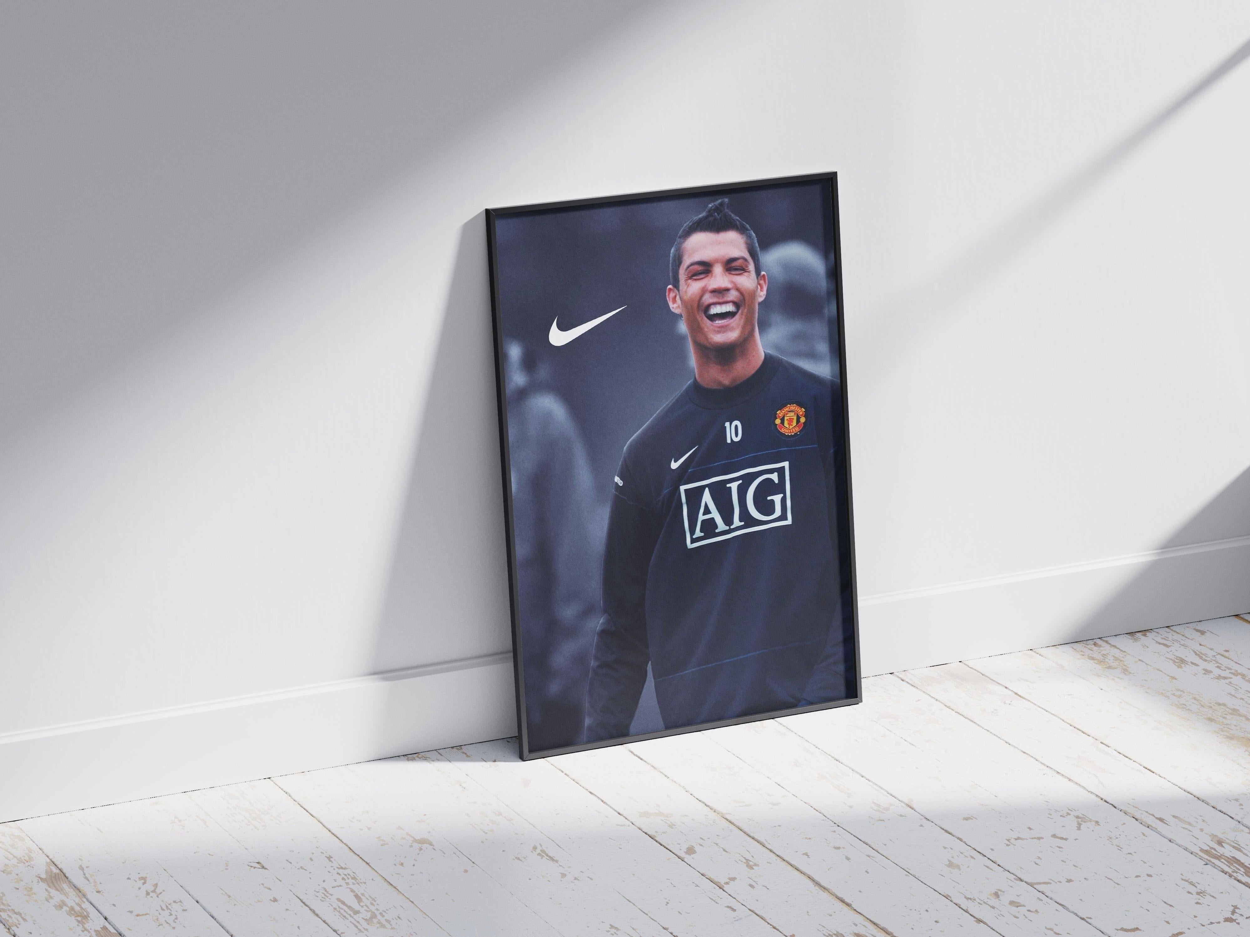 The Cristiano Ronaldo Manchester United Poster by Retro1999 features the star player in a training jersey, smiling, with sponsor logos, framed and leaning against a white wall on a sunlit wooden floor.