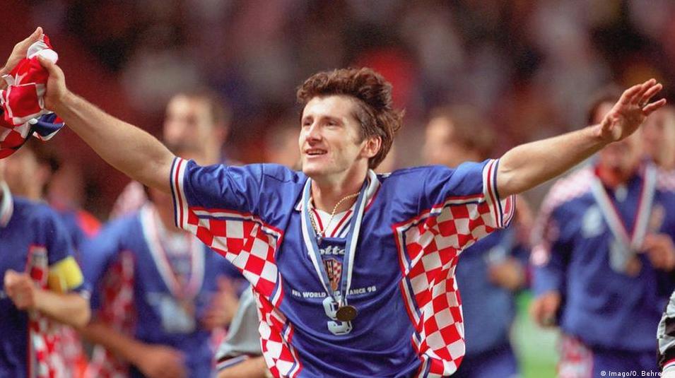 A joyful soccer player in a Retro1999 Croatia 1998 Away Shirt celebrates with arms wide open, medal on neck, and teammates nearby on a blurred stadium field, capturing classic Croatian football spirit.