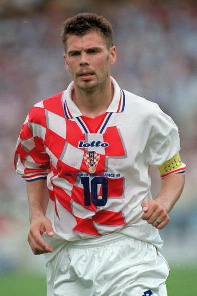 A male soccer player in a Retro1999 Croatia 1998 Home Shirt with red and white checks, number 10, and a captain’s armband runs on the field during a World Cup 1998 match.