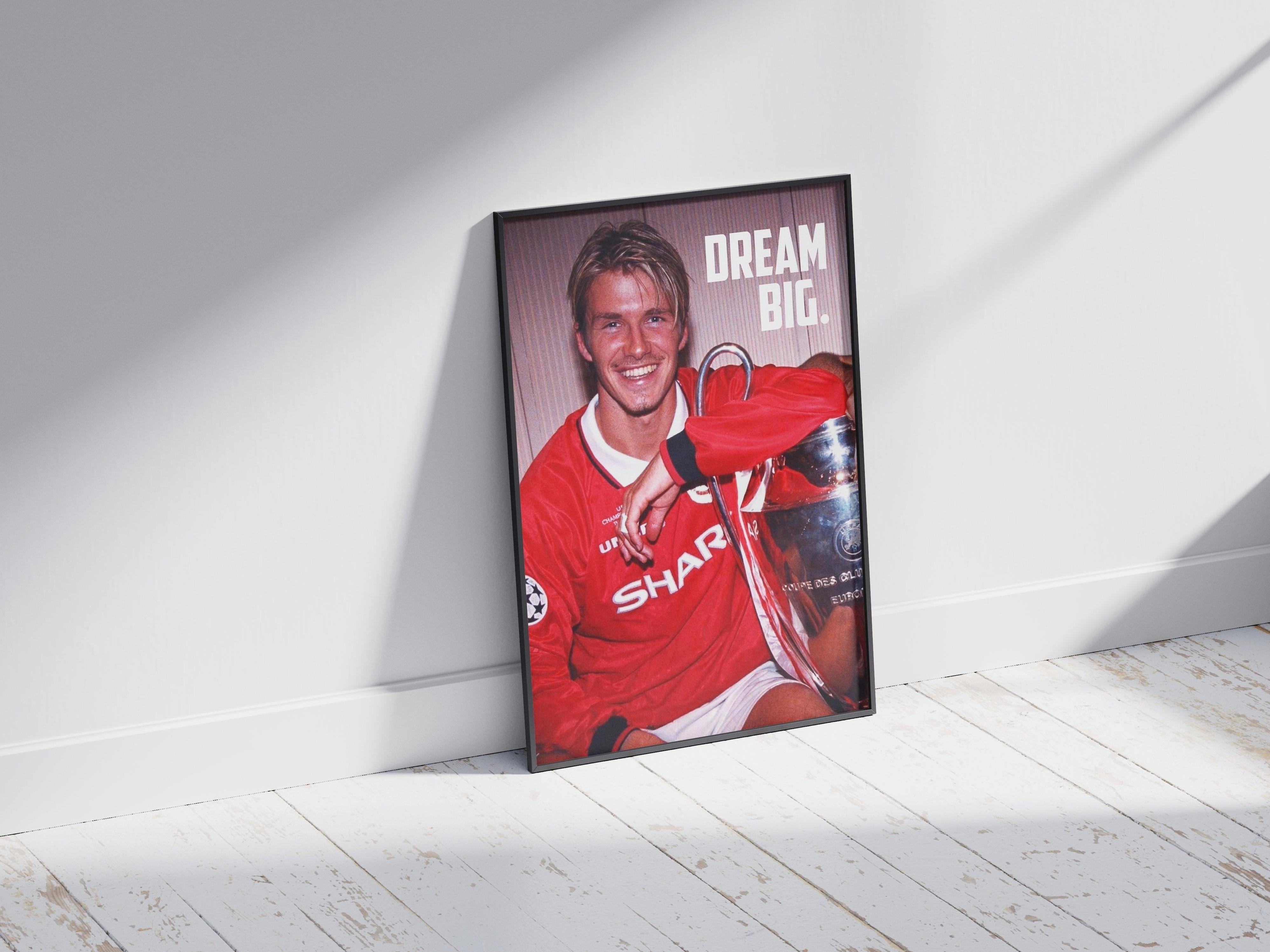The David Beckham Dream Big Poster by Retro1999 features a framed photo of Beckham in a red jersey holding the UEFA Champions League trophy, leaning against a white wall. The words DREAM BIG. appear in the upper right, inspiring all football fans.