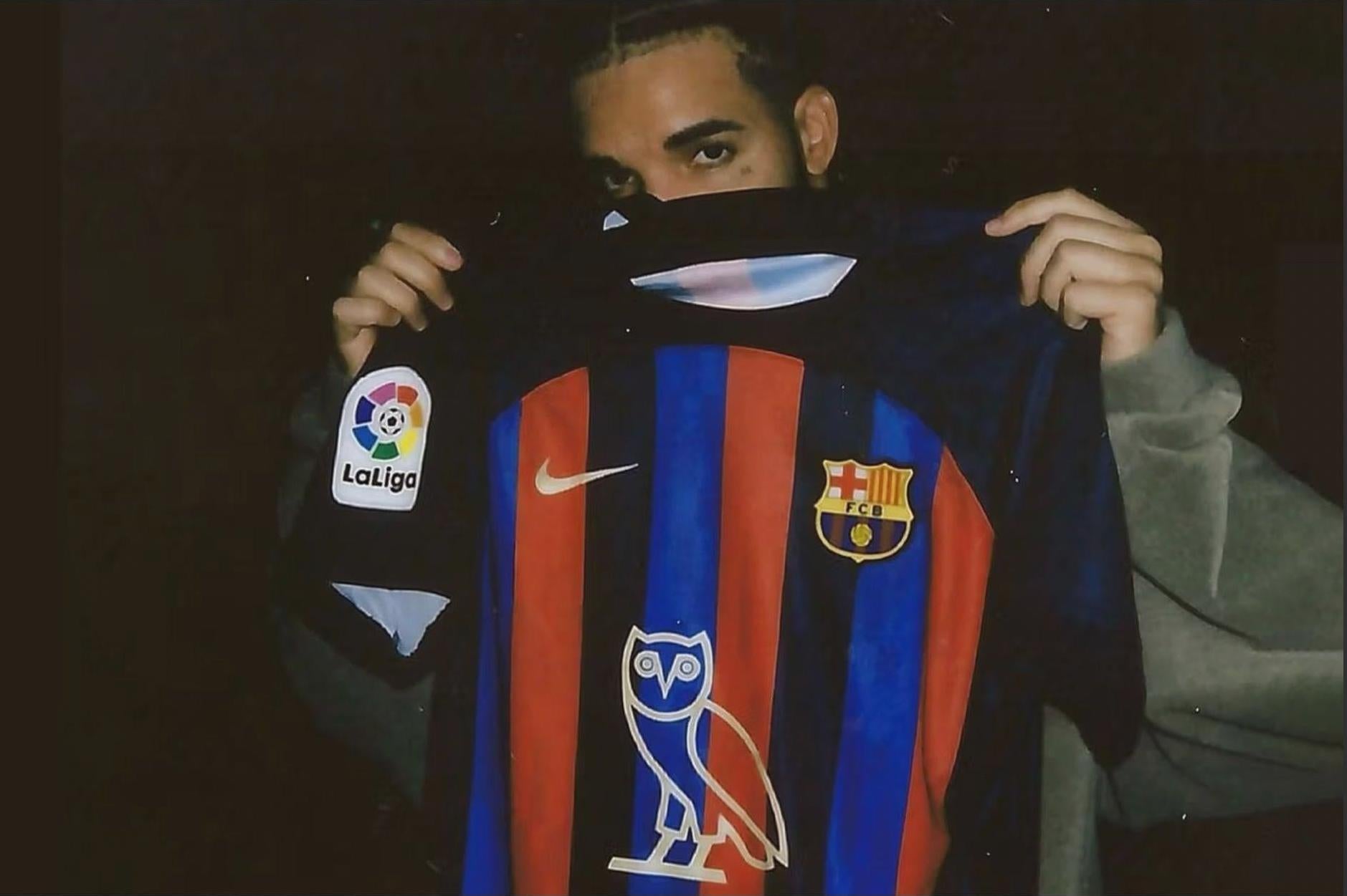 A person holds up the Retro1999 FC Barcelona x Drake - OVO Limited Edition 2022-23 Home Shirt, featuring the La Liga patch, Nike logo, club crest, and OVO owl logo. The dark background partially obscures the person’s face with the shirt.