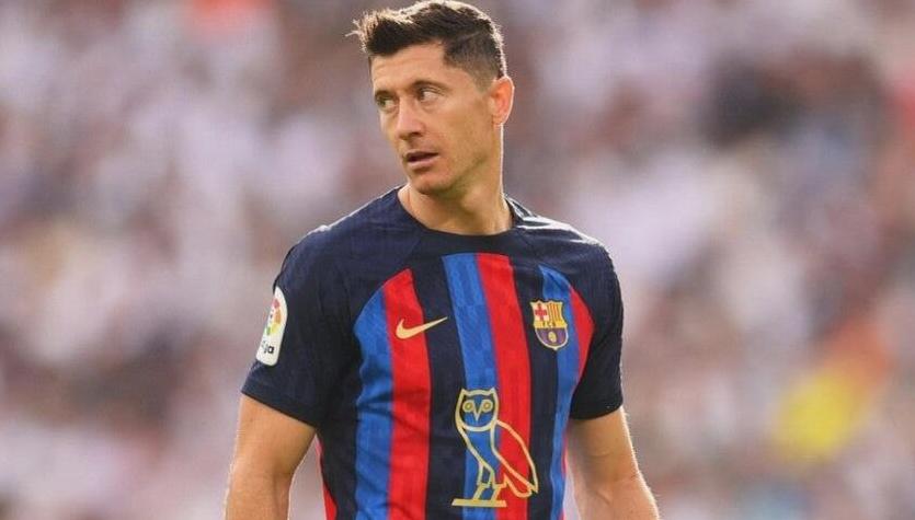 A male soccer player wearing the Retro1999 FC Barcelona x Drake - OVO Limited Edition 2022-23 Home Shirt with blue and red stripes stands on the field, looking left, with a blurred crowd in the background.