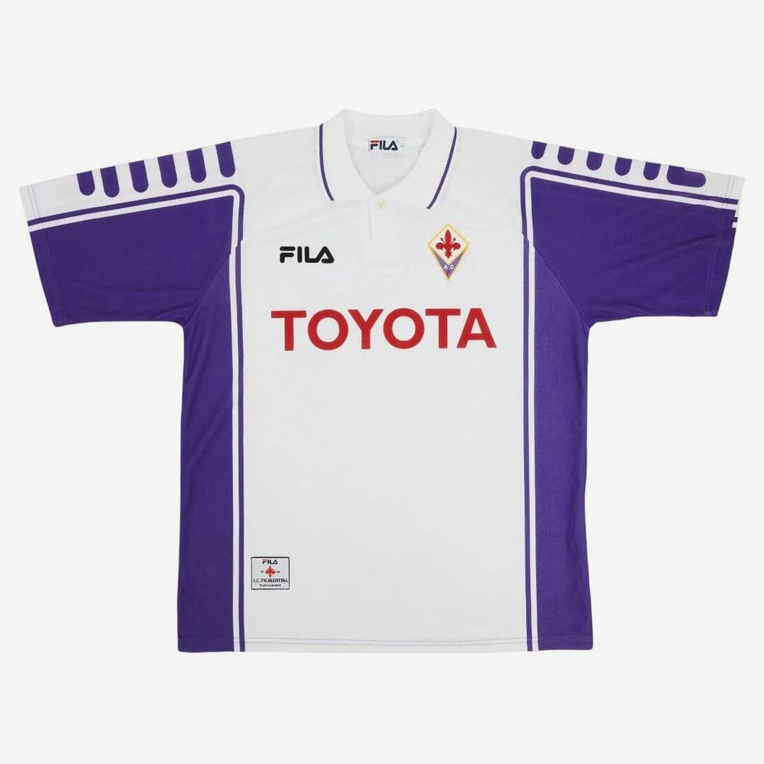 The Retro1999 Fiorentina 1999-00 Away Shirt features a vintage white and purple design with red TOYOTA chest print, FILA logos, bold purple side panels, and the Fiorentina badge—a true retro football classic.