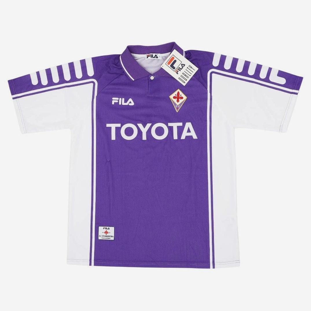The Retro1999 Fiorentina 1999-00 Home Shirt features a purple and white design, TOYOTA sponsor, FILA logo, and club badge on the chest, styled after the iconic retro kit with tag attached to the collar.