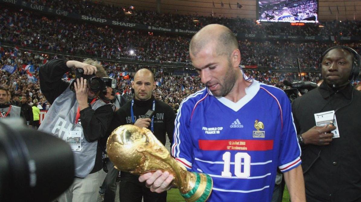 A soccer player wearing the Retro1999 France 1998 Home Shirt with number 10 holds the FIFA World Cup trophy on the field, surrounded by photographers and cameramen, with a cheering crowd in the background.