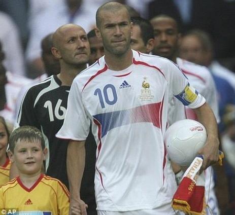 A male soccer player wears the Retro1999 France 2006 Away Shirt with number 10 and captains armband, holding a ball as he walks onto the field with a child and teammates—an iconic football moment.