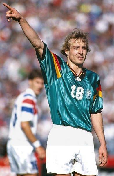 Wearing the Retro1999 Germany 1994 Away Shirt, number 18 raises his right arm and points during a match, with a player in a white and blue kit blurred in the background.