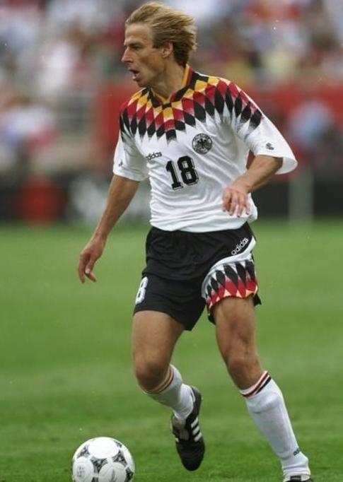A soccer player in the Retro1999 Germany 1994 Home Shirt, number 18, dribbles a ball on grass during a match. Blurred fans in the stands evoke the legacy of German football.