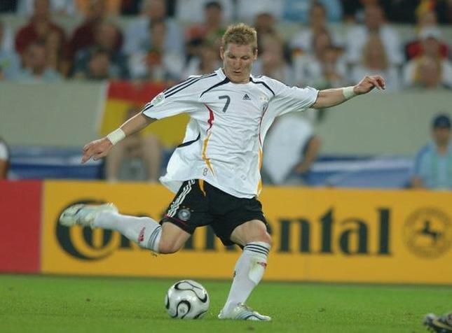 A soccer player in a white Retro1999 Germany 2006 Home Shirt with the number 7 is kicking a ball during a match, with spectators and advertising boards in the background.