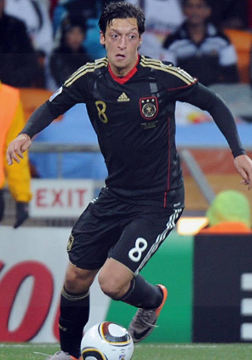 A soccer player for Germany controls the ball on the field wearing a Retro1999 Germany 2010 Away Shirt, number 8. Blurred crowds and teammates are visible in the background.
