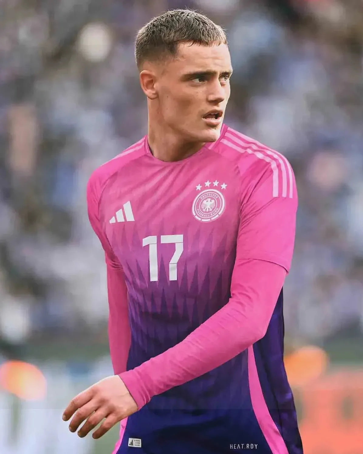 A male soccer player in the Retro1999 Germany 2024 Away Shirt with number 17 stands on the field, gazing right. His retro kit offers iconic style and comfort, set against a blurred stadium packed with spectators.