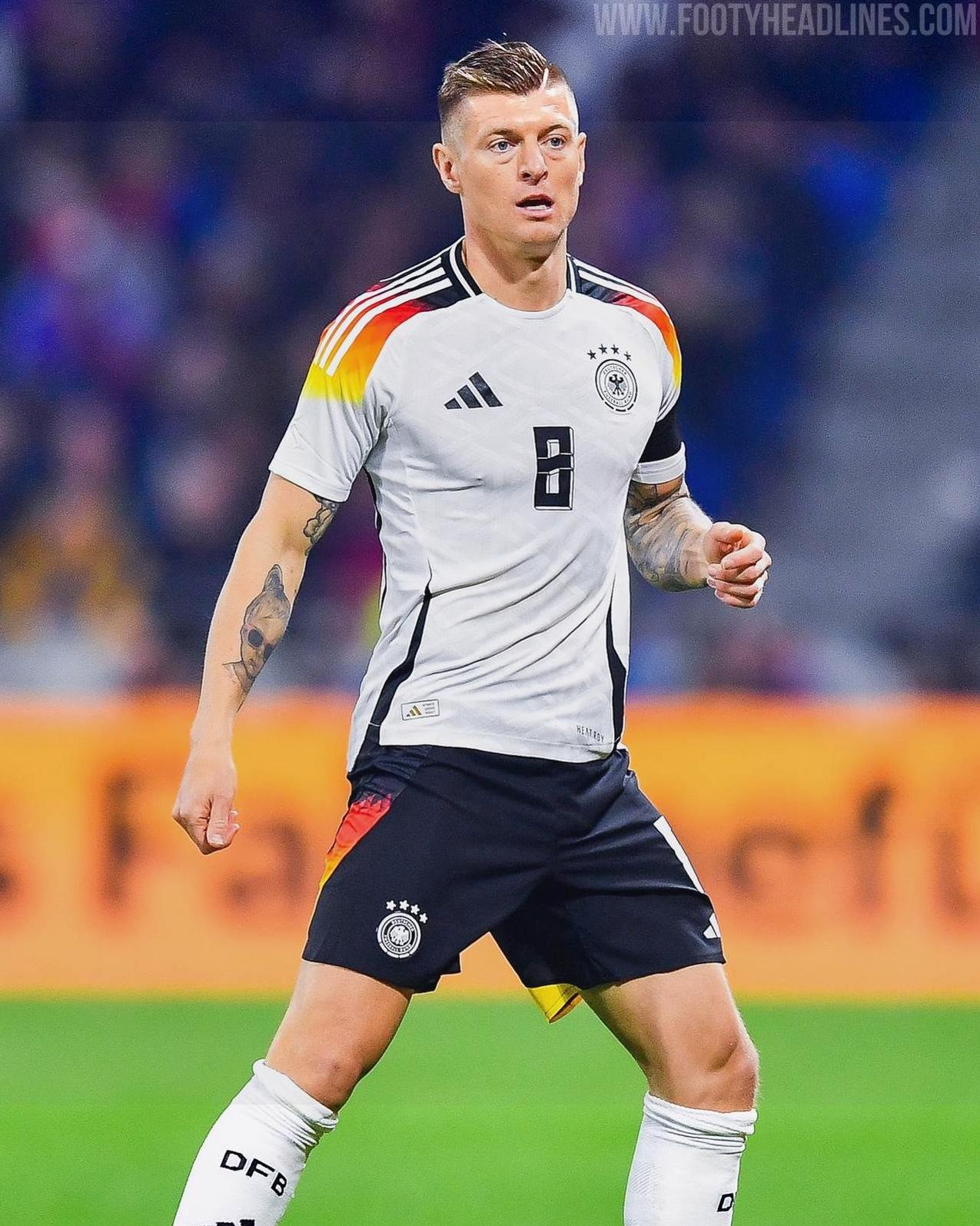 A male soccer player wearing the Retro1999 Germany 2024 Home Shirt with black shorts is on the field. The shirt has red, yellow, and black shoulder details, and he has visible tattoos on his left arm.