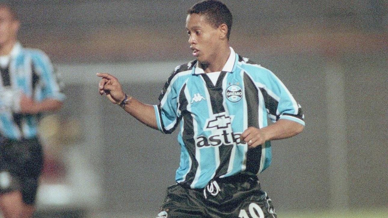 A young male soccer player in a Retro1999 Grêmio 1999-00 Home Shirt runs on the field, focused and pointing with his right arm—channeling classic Ronaldinho Gaúcho retro style.