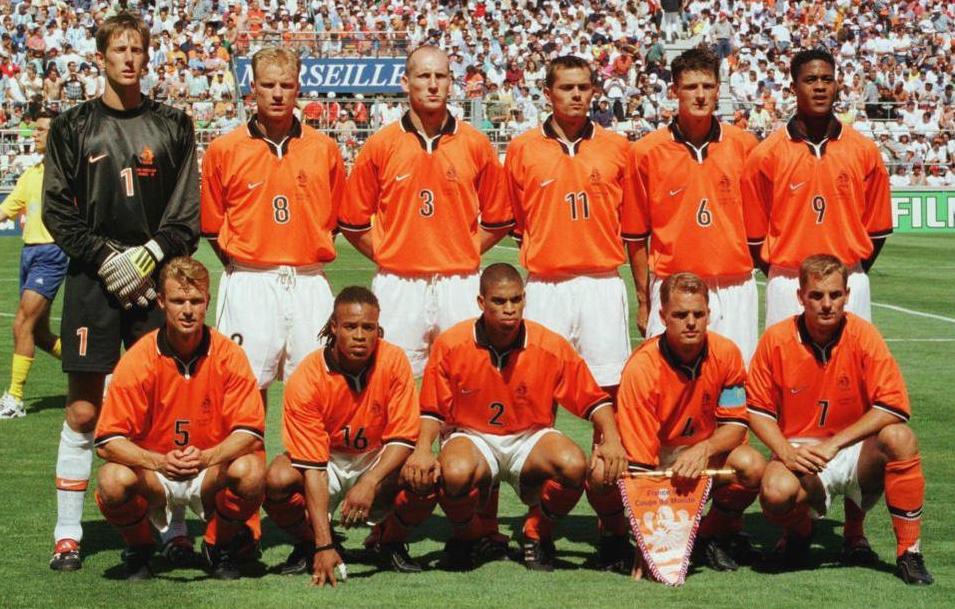 Eleven male soccer players wear the Retro1999 Holland 1998 Home Shirt as they pose for a team photo on a grassy field, with fans in the stadium behind them. The Dutch squad is arranged in two rows, standing and kneeling.