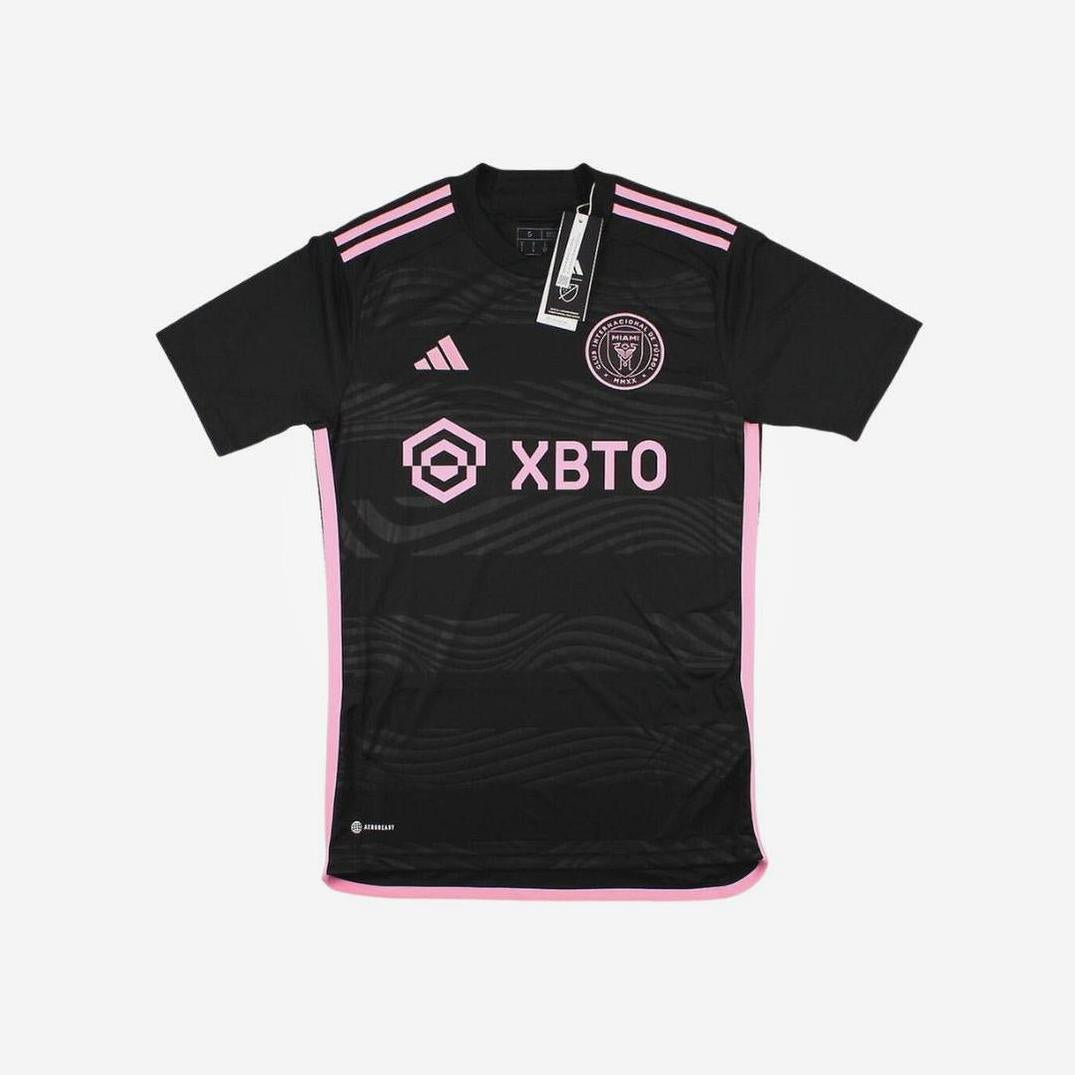 The Retro1999 Inter Miami 2023/24 Away Shirt is black with pink accents, XBTO sponsor logo, club crest on the chest, pink shoulder stripes, and a collar tag. Worn by stars like Messi during MLS matches.