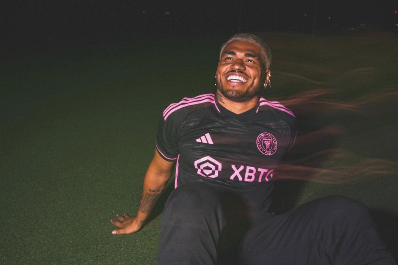A smiling person with bleached hair sits on green turf at night, wearing the Retro1999 Inter Miami 2023/24 Away Shirt. Light trails streak beside them, evoking the dynamic spirit of Lionel Messi’s team.