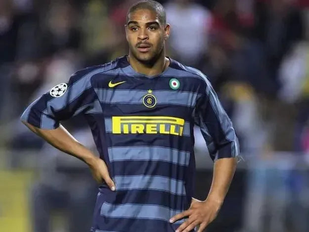 A football player wearing the Retro1999 Inter Milan 2004-05 Third Shirt, featuring blue and gray stripes, stands with his hands on his hips on the field during a match.