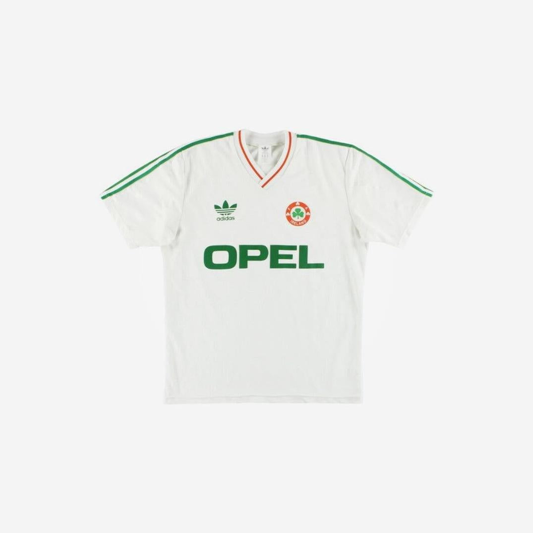 The Ireland 1990 Away Shirt by Retro1999 is a vintage white soccer jersey with green Adidas stripes, a green OPEL sponsor, an orange-and-green crest, and a collar trimmed in red and green—a classic tribute to football heritage.
