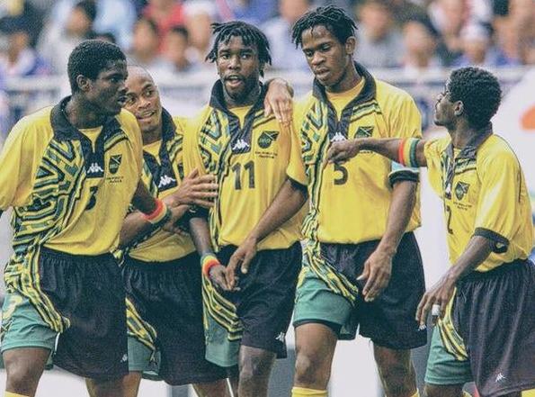 Five players in Retro1999’s Jamaica 1998 Home Shirt celebrate on the field, one Reggae Boyz star gesturing energetically, with a blurred crowd in the background.
