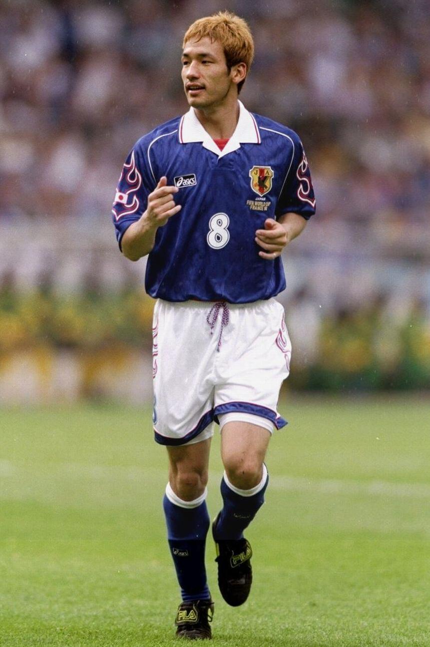 A soccer player in a Retro1999 Japan 1998 Home Shirt with the number 8 runs on a grassy field during a match, with a blurred crowd in the background, reflecting the passion of Japanese football.