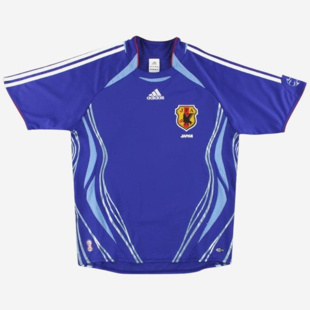 The Japan 2006 Home Shirt by Retro1999 features a blue design with white and light blue accents, short sleeves with three white stripes, central Adidas logo, and team crest on the left chest—celebrating Japanese football history.
