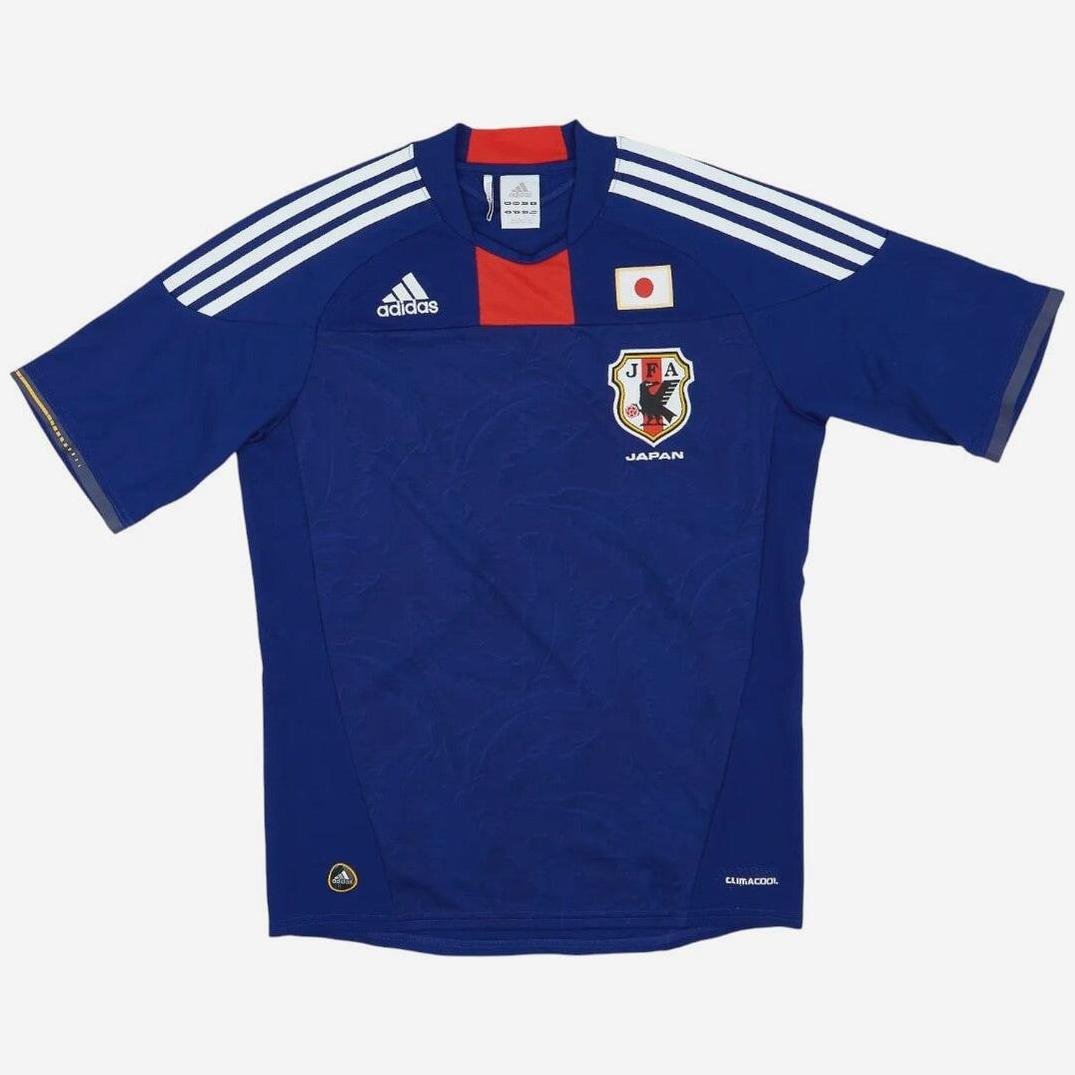 The Japan 2010 Home Shirt by Retro1999 features blue fabric, white shoulder stripes, red collar detail, Adidas branding, the Japan flag, and team crest—a classic football kit celebrating legacy.